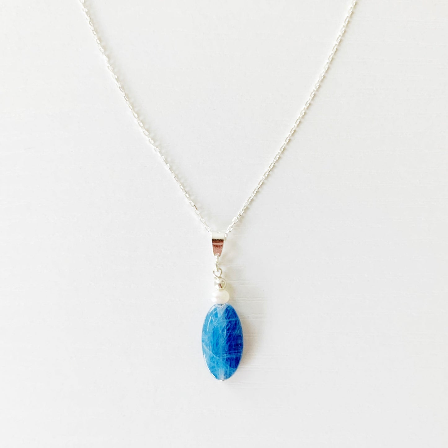 the brewster necklace by mermaids and madeleines is a pendant style necklace with a long oval blue kyanite bead with a pearl on top dropped from a sterling silver chain with sterling findings. this necklace is photographed flat on a white surface