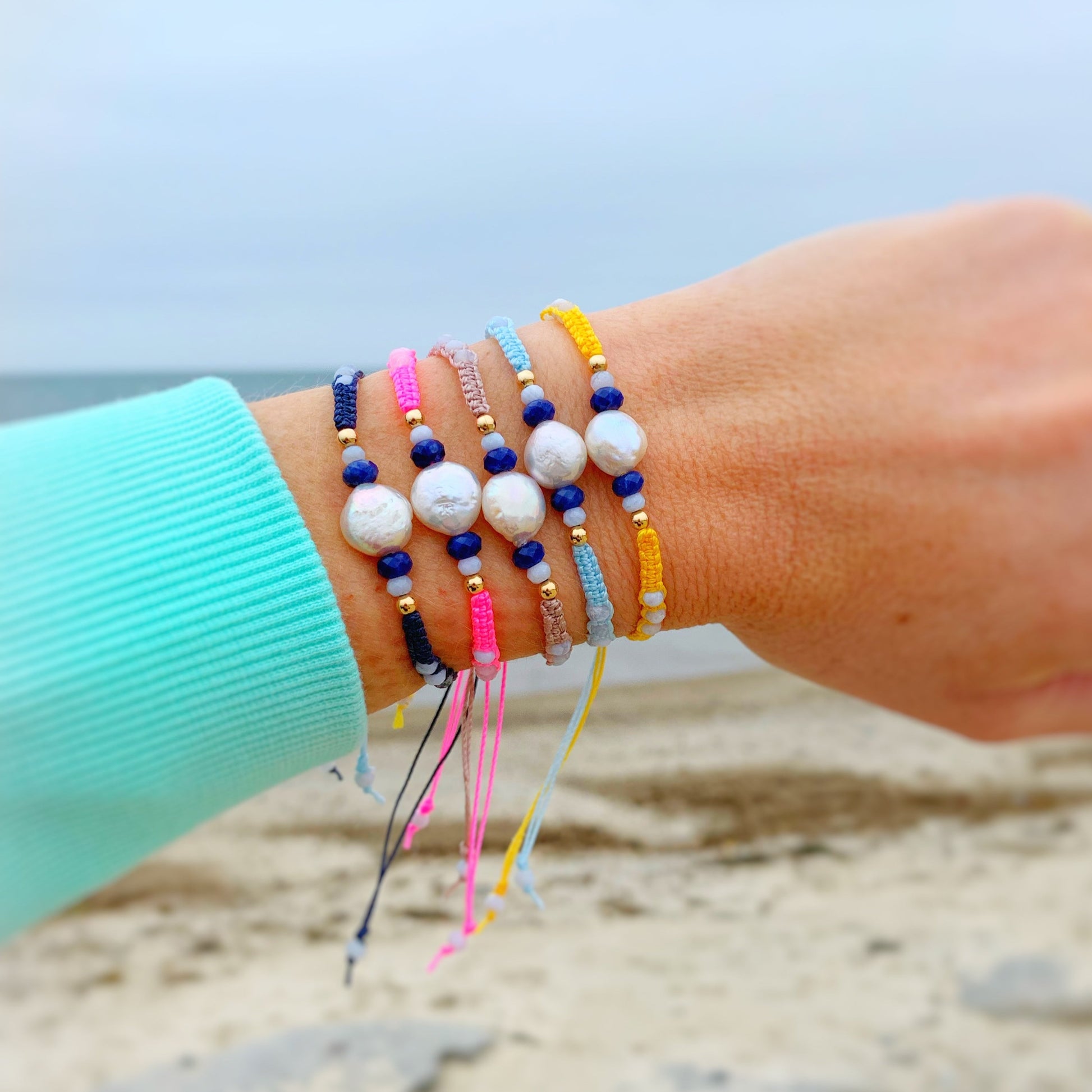 the 5 available colors of the bristol macrame friendship bracelets by mermaids and madeleines stacked up and worn on a wrist with the ocean in the backdrop of the photo