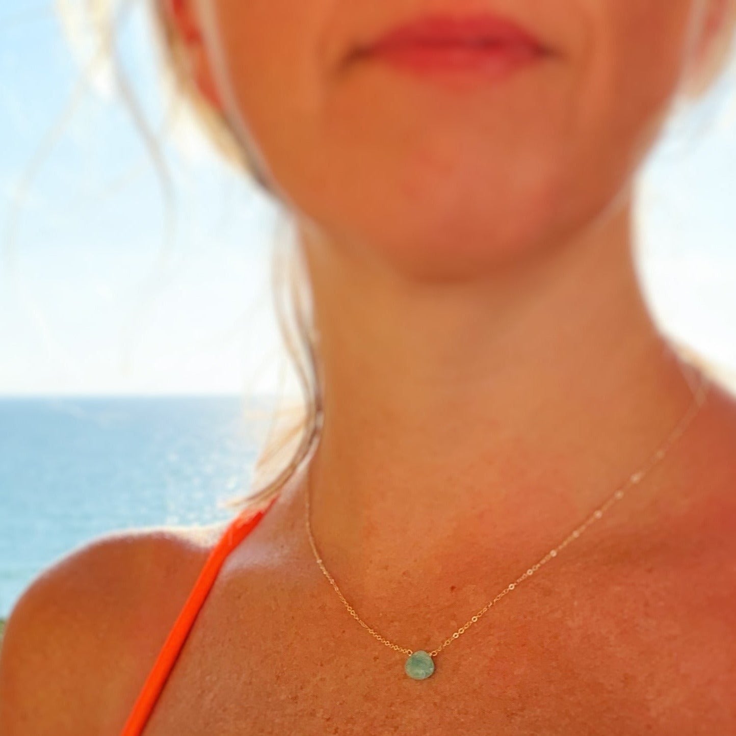 The captiva necklace by mermaids and madeleines is a light weight necklace created with a bright aqua amzonite faceted briolette at the center of 14k gold filled chain. this necklace is photographed close up on a person with a blurry ocean background