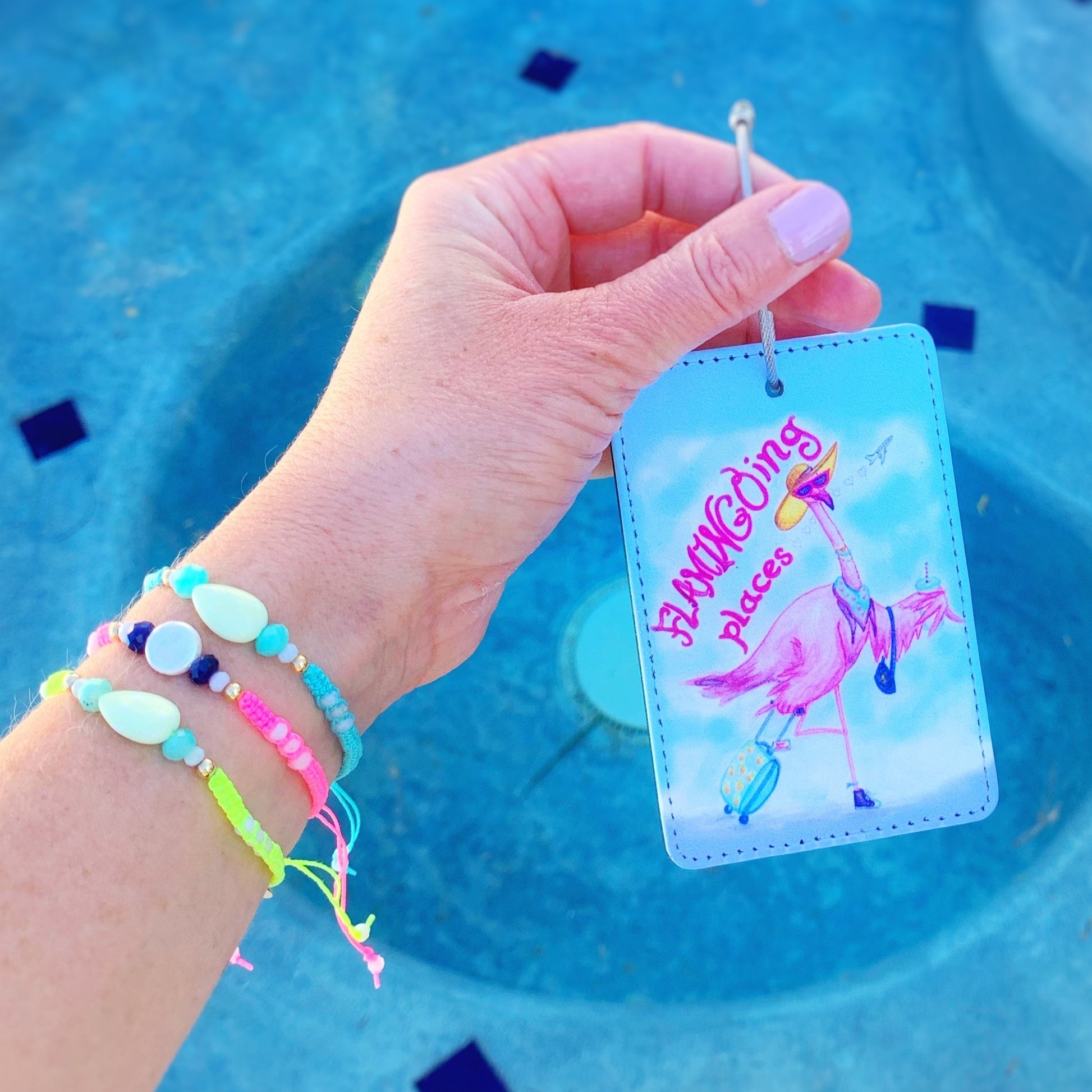 the flamingal luggage tag has a flamingo wearing a hat and sunglasses carring a suitcase on it. this tag is photographed held up in a hand with a blue pool in the background