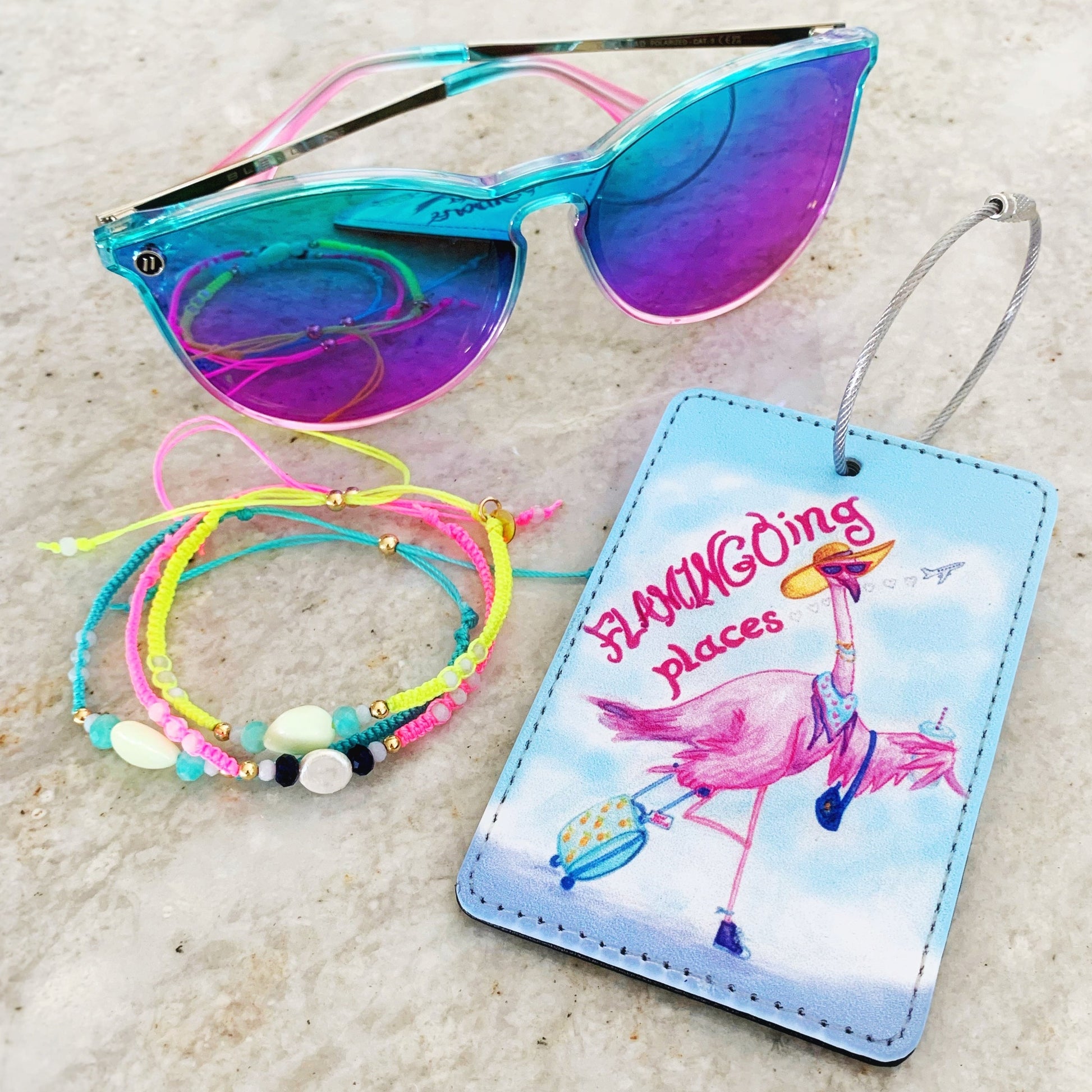 the flamingal luggage tag has a flamingo on it wearing a hat and sunglasses and wheeling a suitcase behind. this tag is photographed flat on a surface next to a pair of sunglasses and a stack of macrame bracelets