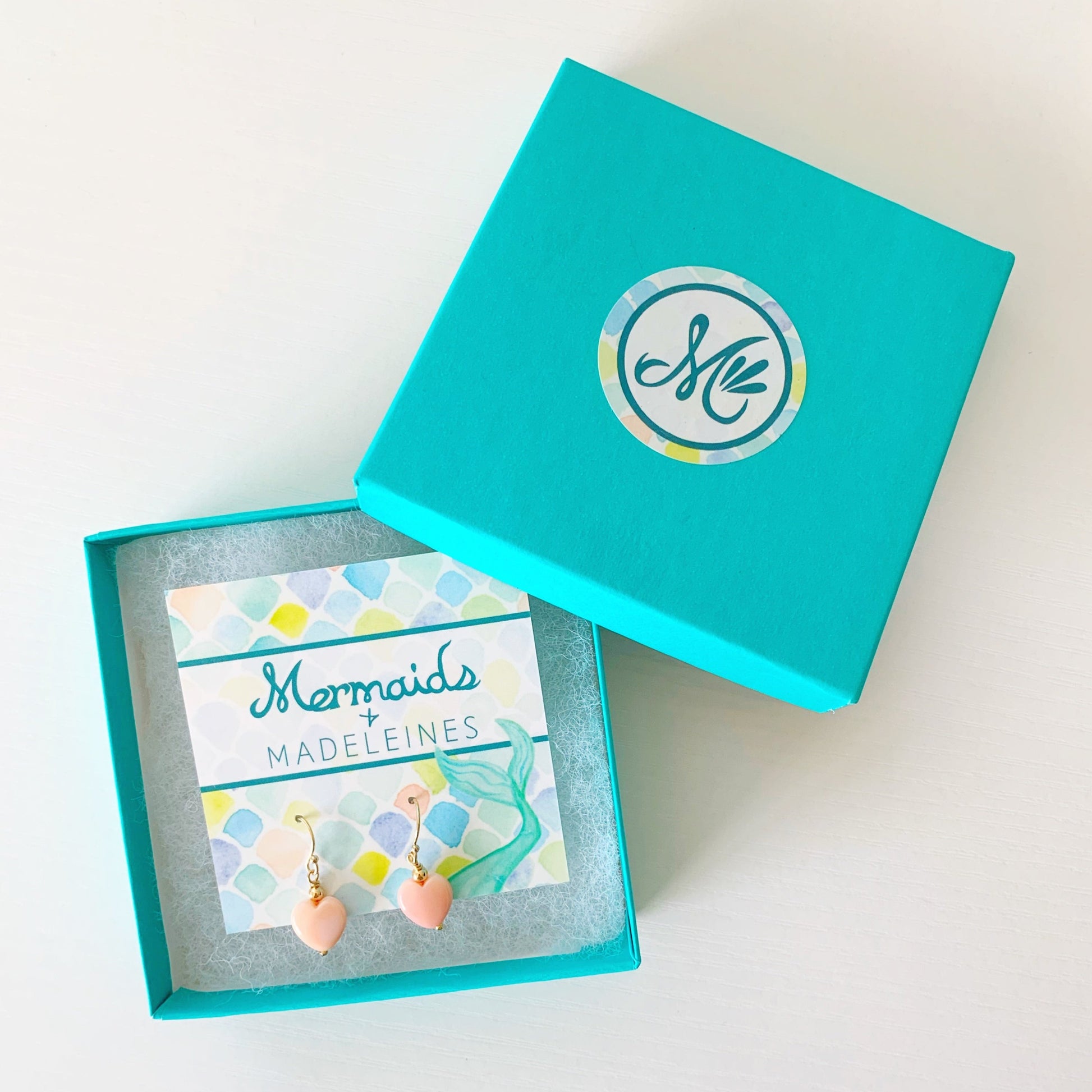 the pink posy earrings by mermaids and madeleines are simple drop earrings created with 14k gold beads and findings and feature a conch shells carved into hearts. this pair is carded and photographed in a teal gift box on a white surface