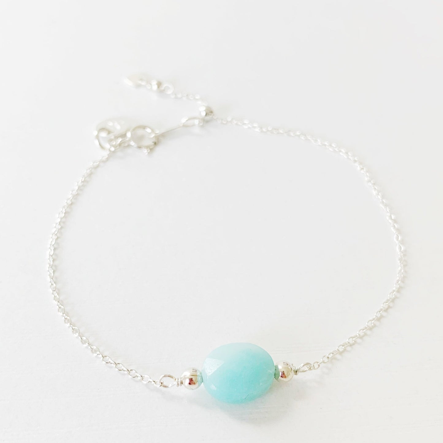 the mermaids and madeleines bracelet is a sterling silver chain bracelet with a faceted amazonite oval shaped bead at the center and a slide bead near the clasp to make it adjustable. this bracelet is photographed on a white surface