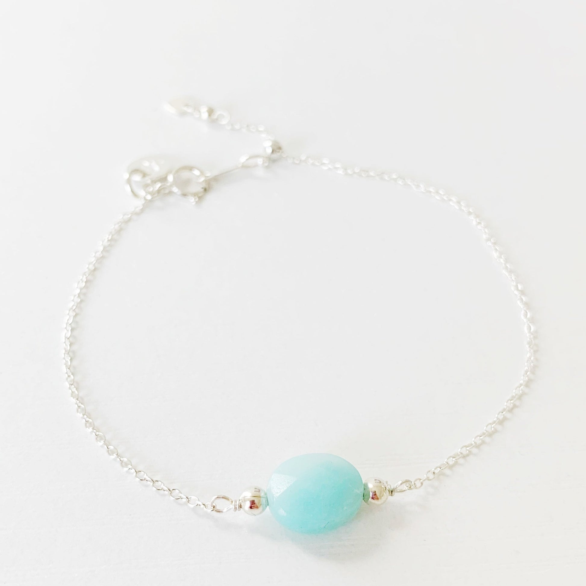 the mermaids and madeleines bracelet is a sterling silver chain bracelet with a faceted amazonite oval shaped bead at the center and a slide bead near the clasp to make it adjustable. this bracelet is photographed on a white surface