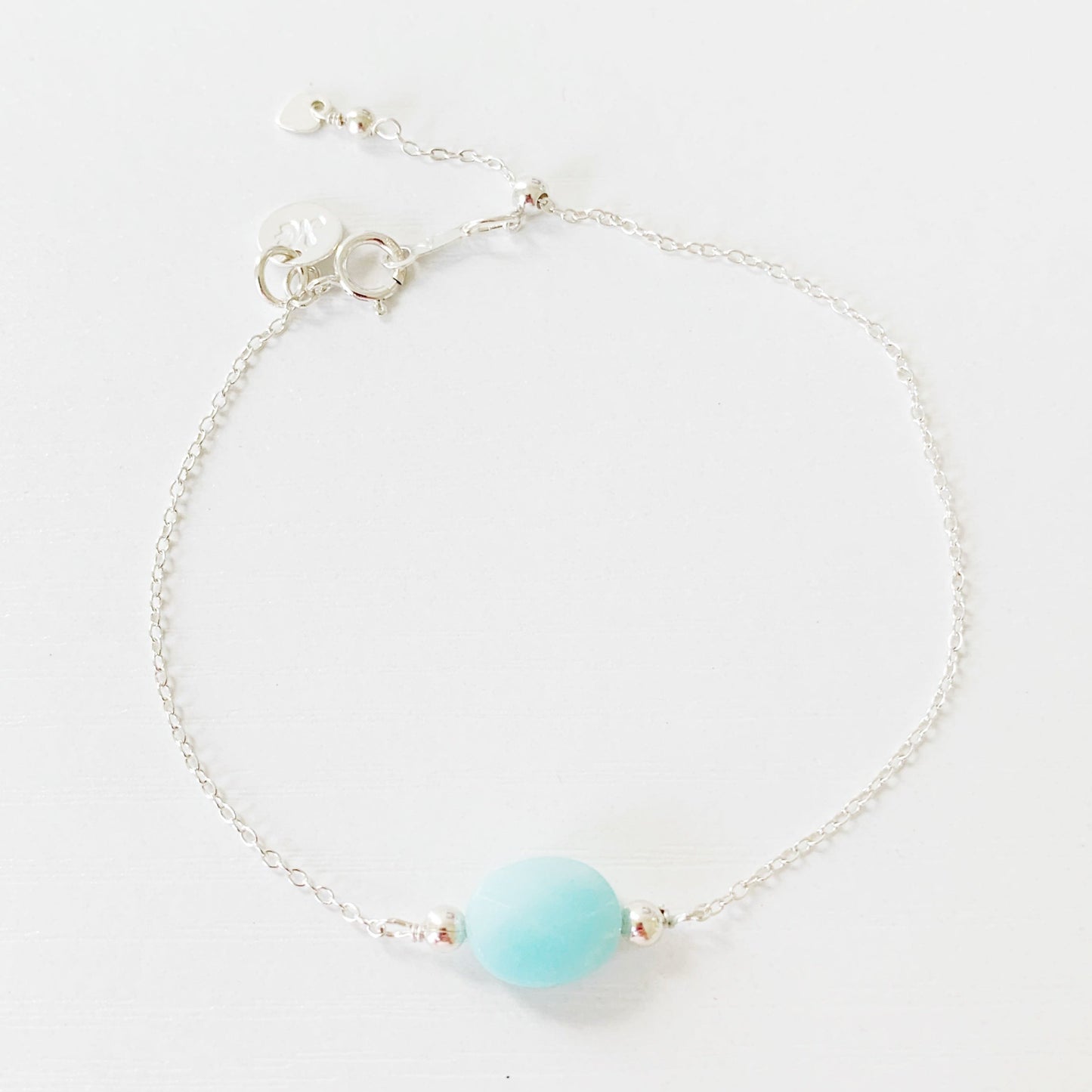 the mermaids and madeleines bracelet is a sterling silver chain bracelet with a faceted amazonite oval shaped bead at the center and a slide bead near the clasp to make it adjustable. this bracelet is photographed on a white surface