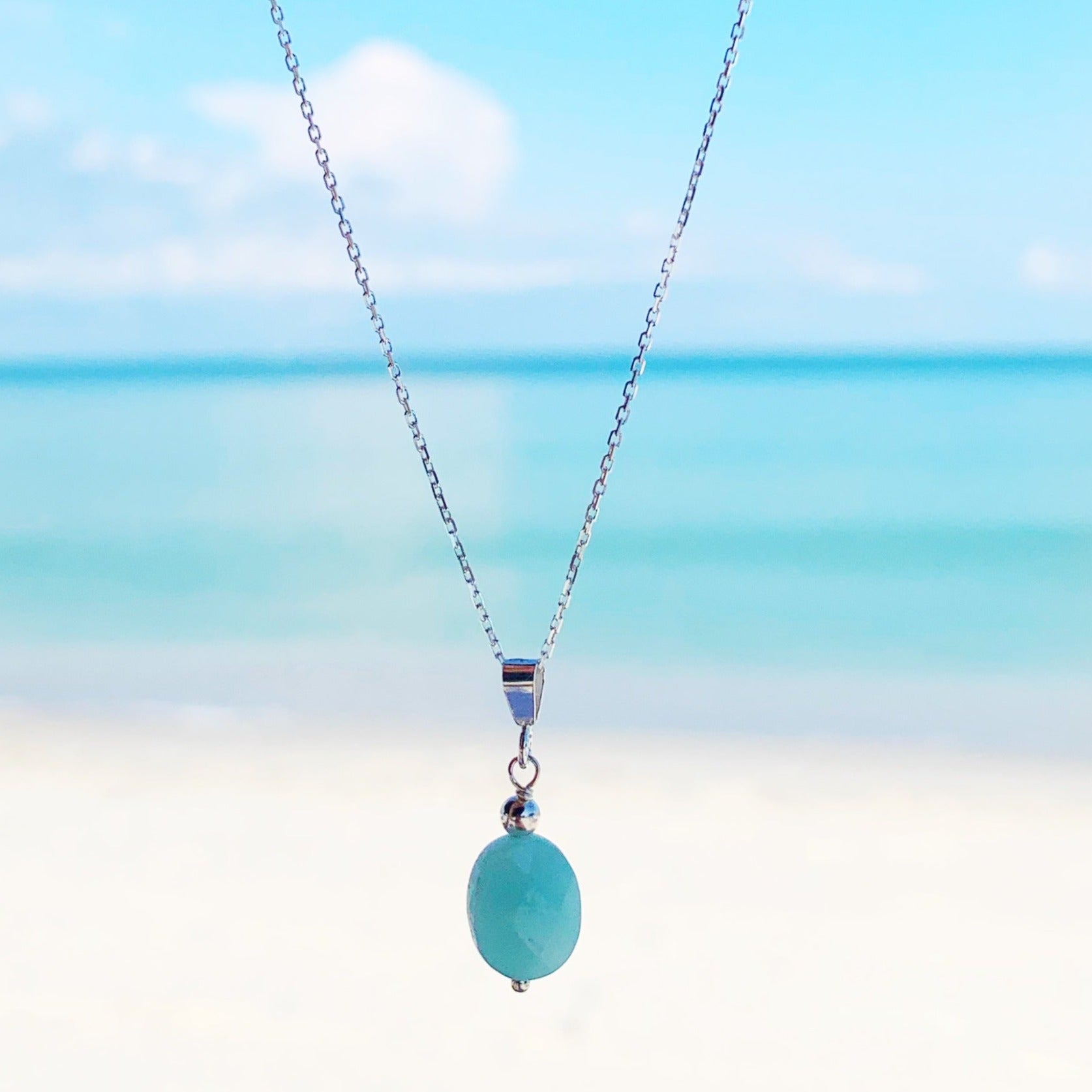 the laguna necklace by mermaids and madeleines is a faceted oval amazonite bead suspended from sterling silver chain and findings. this necklace is photographed closer up with a hazy beach background