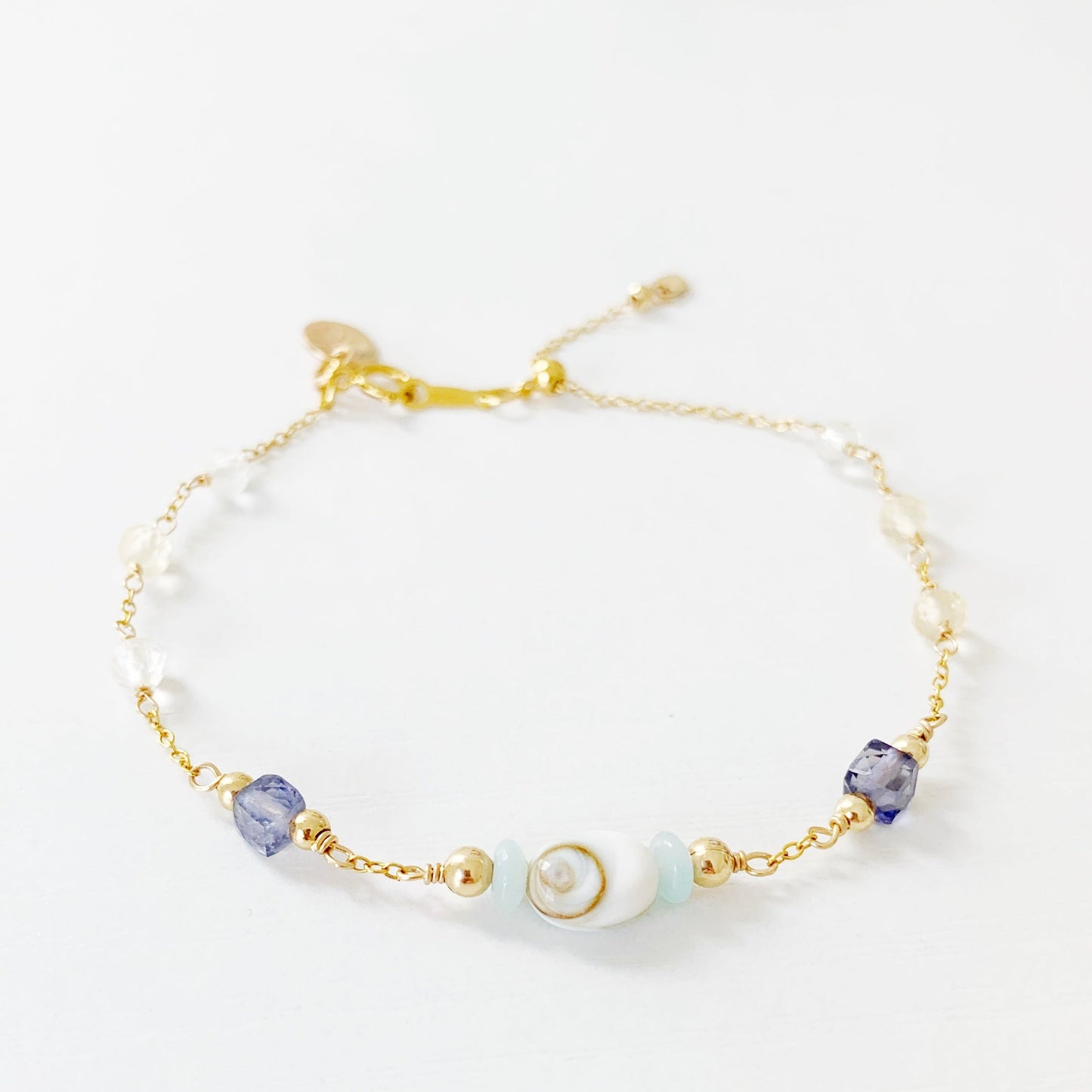 mermaid money adjustable bracelet by mermaids and madeleines is a 14k gold filled chain bracelet with beads throughout and a shiva shell at the center. theres a slide bead near the clasp. the bracelet is photographed on a white surface