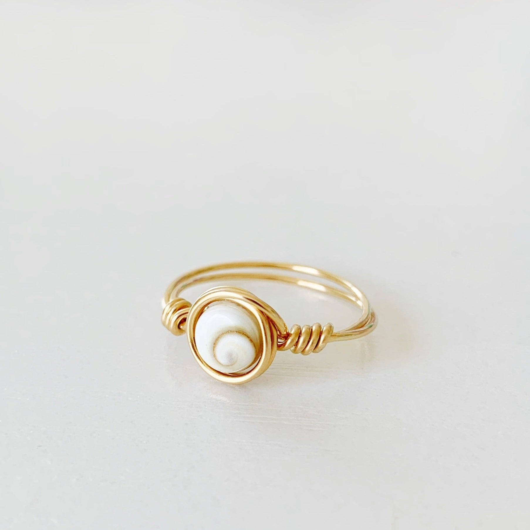 the mermaid money ring by mermaids and madeleines is a wire wrapped ring with a coin shaped shiva shell bead at the center with 14k gold filled wire swirled around it to form a ring. this ring is photographed flat on a white surface