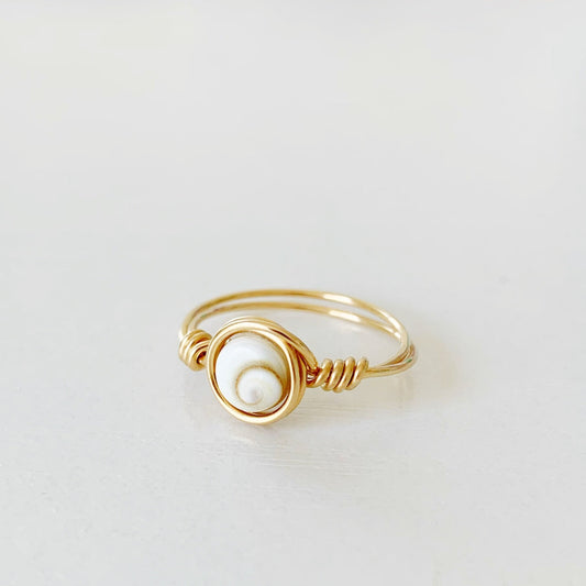 the mermaid money ring by mermaids and madeleines is a wire wrapped ring with a coin shaped shiva shell bead at the center with 14k gold filled wire swirled around it to form a ring. this ring is photographed flat on a white surface