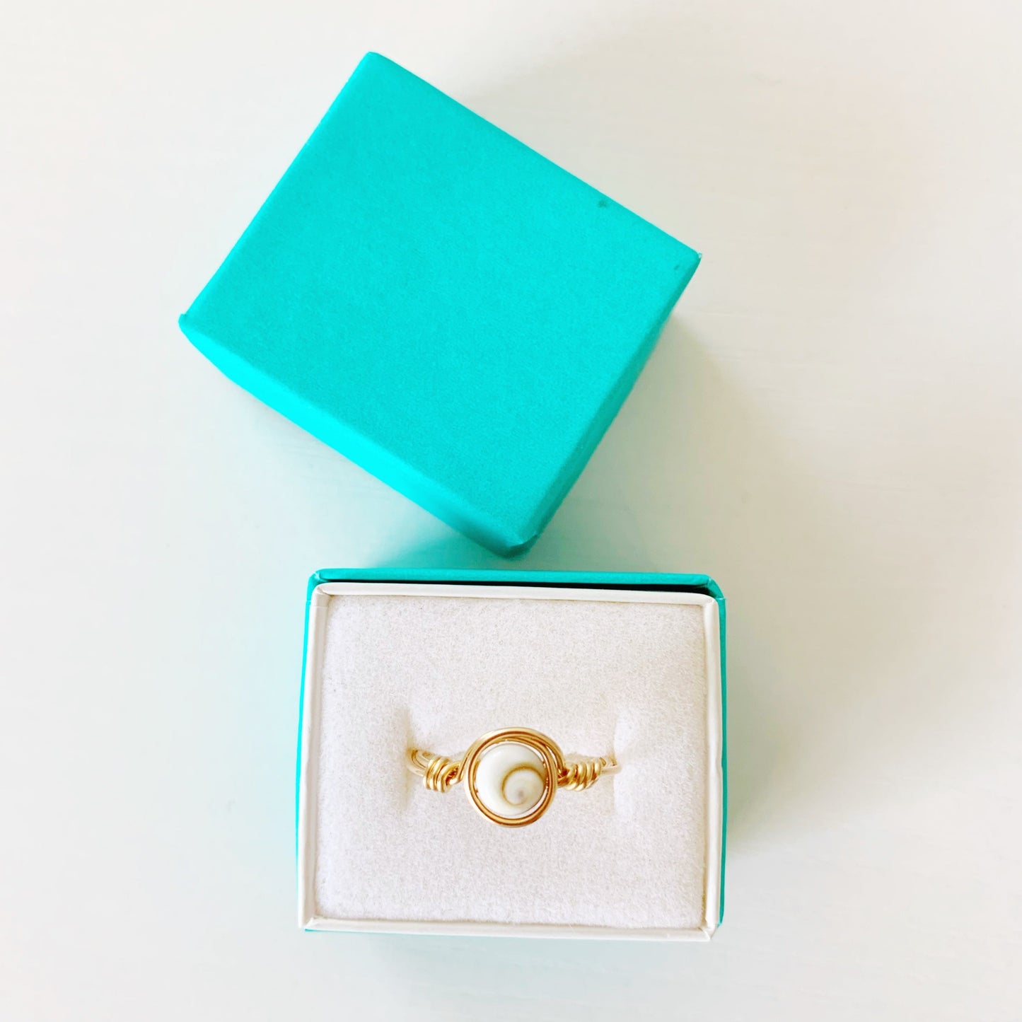 the mermaids and madeleines mermaid money ring is a wire wrapped ring created with a coin shaped shiva shell bead at the center swirled with 14k gold filled wire to form the ring. this ring is photographed in a teal paper lined ring box on a white surface