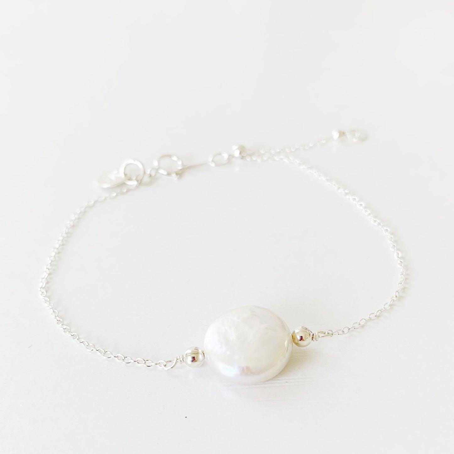 the mermaids and madeleines newport bracelet has a freshwater coin pearl on the center of sterling silver chain with an adjustable slide bead near the clasp. this bracelet is photographed flat on a white surface