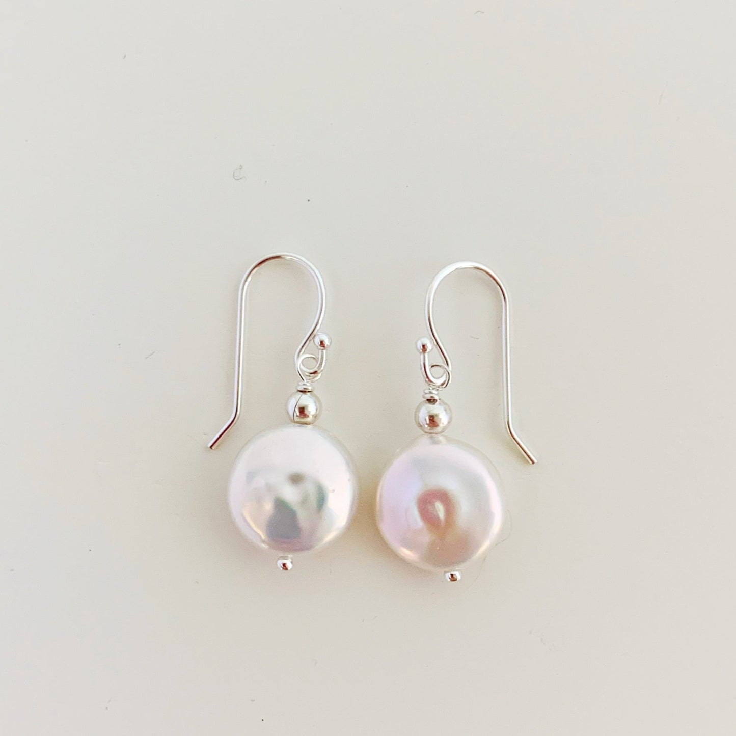 the newport earrings by mermaids and madeleines are a simple style earring designed with white, freshwater coin pearls and sterling silver findings and beads. this pair is photographed flat on a white surface