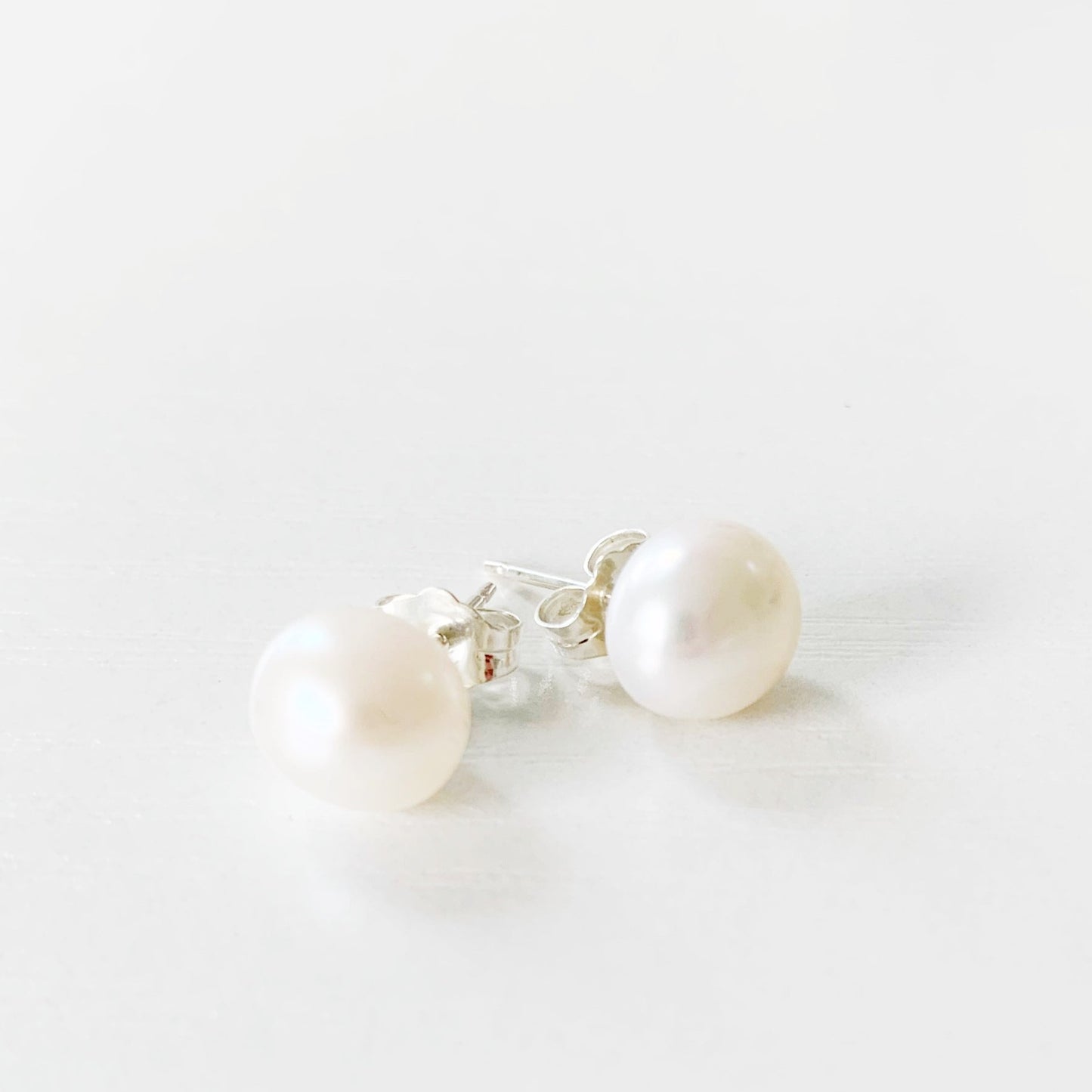 the pearlfect day earrings by mermaids and madeleines are a pair of freshwater button pearl studs with sterling silver findings. this pair is photographed setting on a white surface