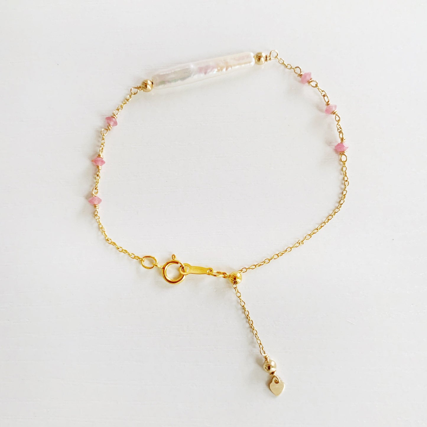 the pink azalea adjustable bracelet by mermaid and madeleines in a 14k gold filled chain bracelet with a slide bead at the clasp. the bracelet features a freshwater pearl bar at the center with pink tourmaline beads going down the chain. this bracelet is photographed flat on a white surface with a top down view