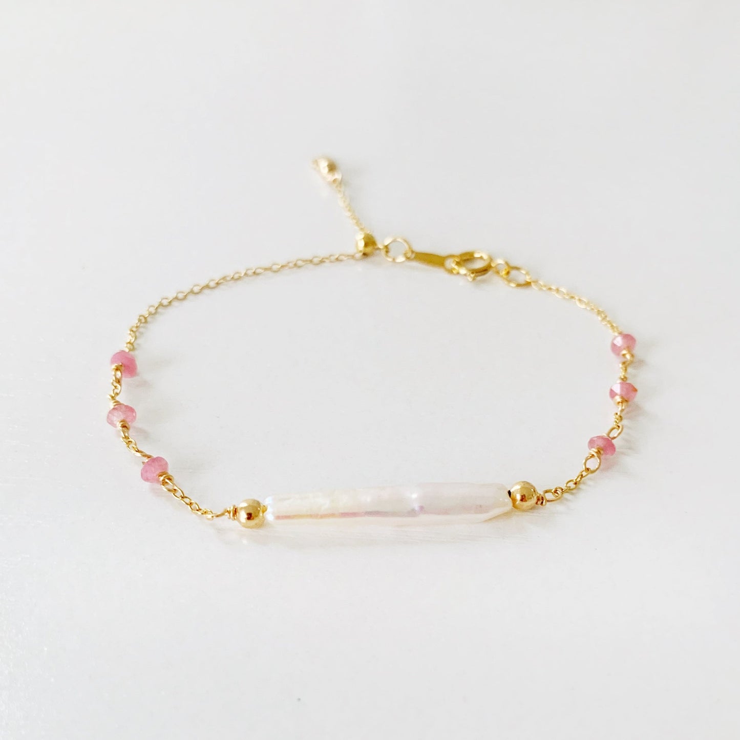 the pink azalea adjustable bracelet by mermaid and madeleines in a 14k gold filled chain bracelet with a slide bead at the clasp. the bracelet features a freshwater pearl bar at the center with pink tourmaline beads going down the chain. this bracelet is photographed flat on a white surface with a front view