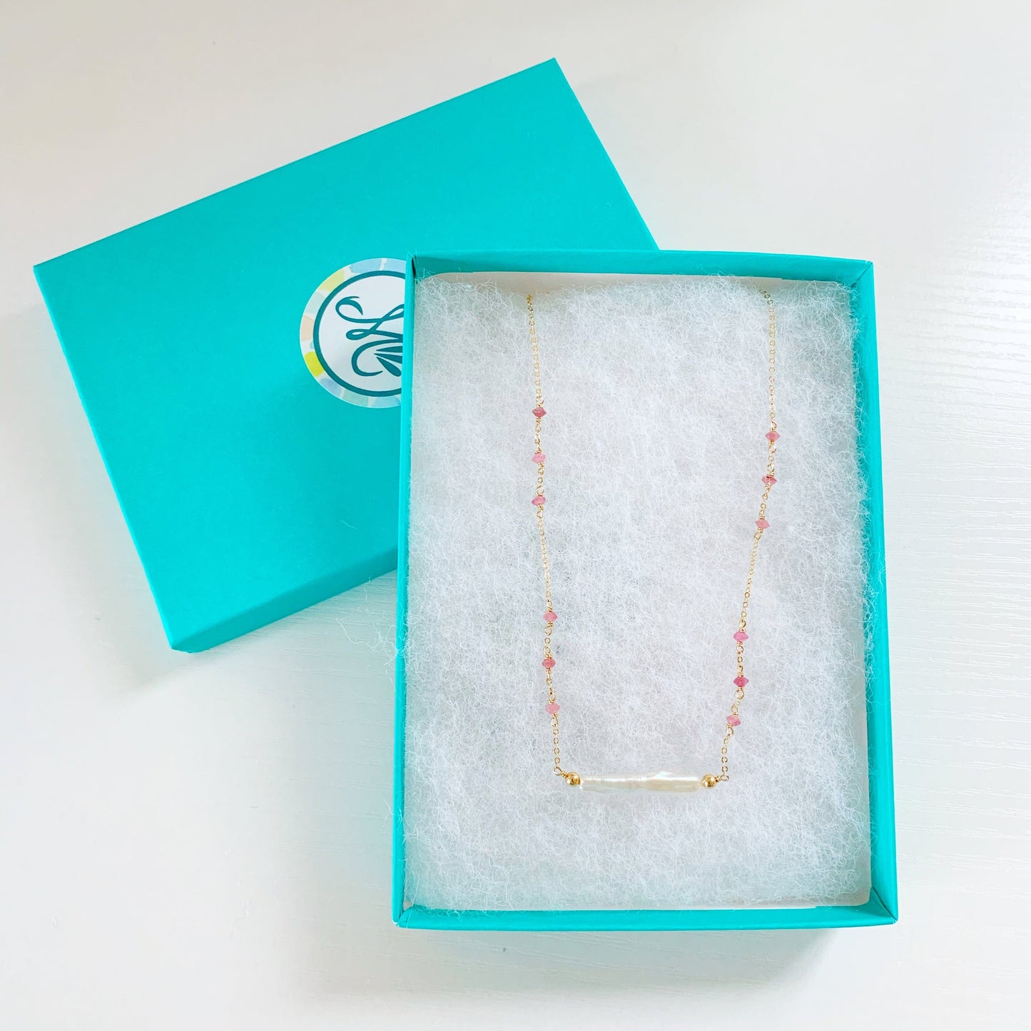 the pink azalea necklace by mermaids and madeleines is a freshwater pearl bar necklace with pink tourmaline faceted beads in groups of 3 on the 14k gold filled chain. this necklace is photographed in a teal gift box on a white surface