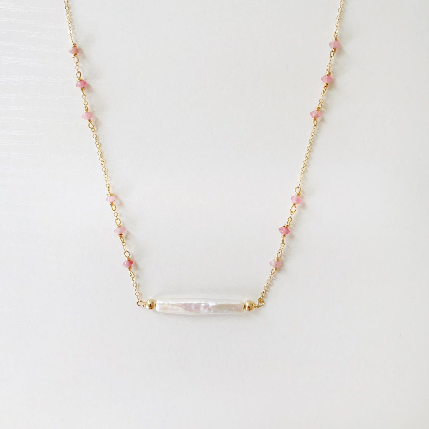 the pink azalea necklace by mermaids and madeleines is a freshwater pearl bar necklace with pink tourmaline faceted beads in groups of 3 on the 14k gold filled chain. this necklace is photographed with the front half in view on a white surface.