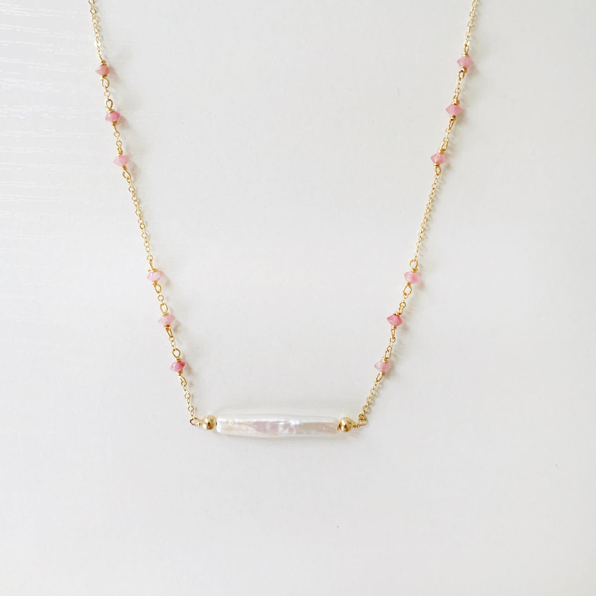 the pink azalea necklace by mermaids and madeleines is a freshwater pearl bar necklace with pink tourmaline faceted beads in groups of 3 on the 14k gold filled chain. this necklace is photographed with the front half in view on a white surface.