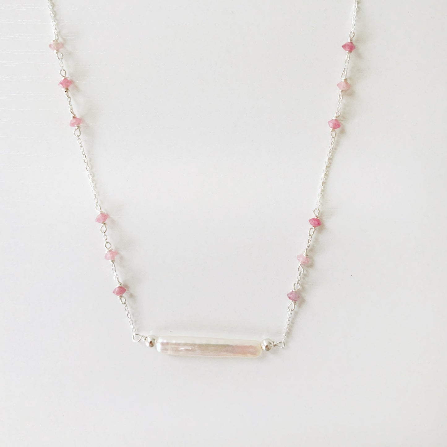 the pink azalea necklace by mermaids and madeleines is a sterling silver chain necklace with a freshwater pearl bar at the center and small pink tourmaline beads in groups of 3 going up the sides of the chain. this necklace is photographed on a white surface with the front half of the necklace in view