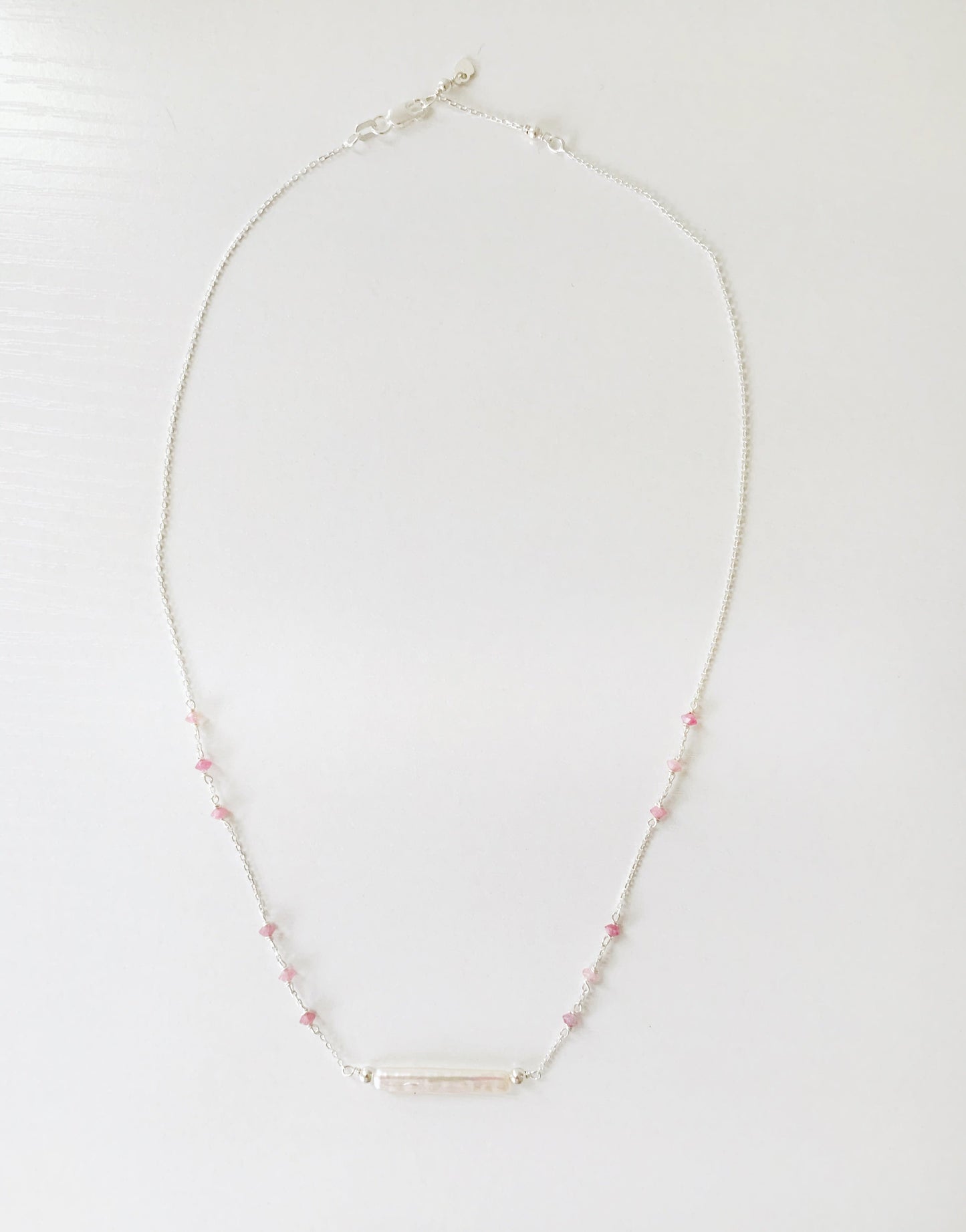 the pink azalea necklace by mermaids and madeleines is a sterling silver chain necklace with a freshwater pearl bar at the center and small pink tourmaline beads in groups of 3 going up the sides of the chain. this necklace is photographed on a white surface with the full length of the necklace in view