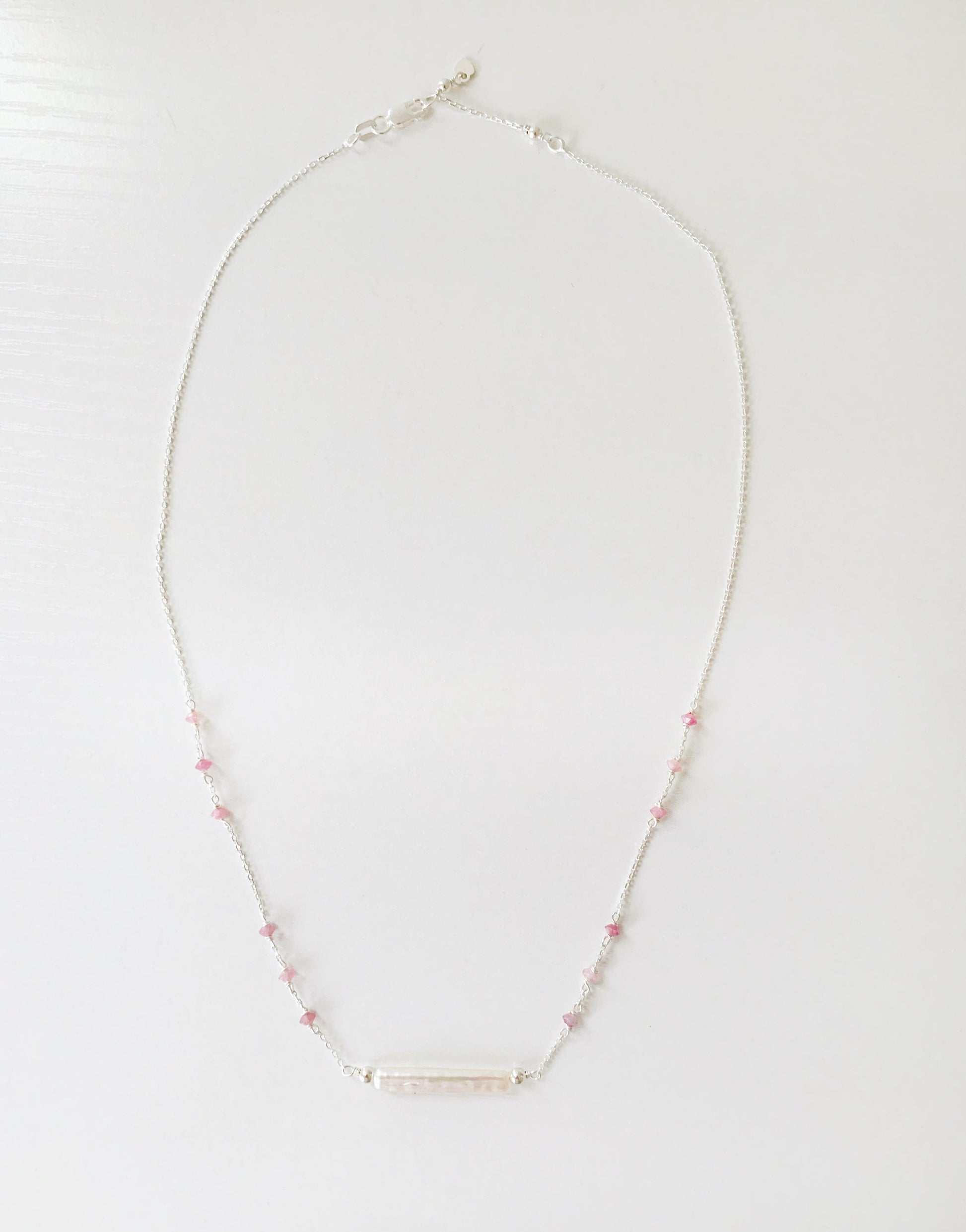 the pink azalea necklace by mermaids and madeleines is a sterling silver chain necklace with a freshwater pearl bar at the center and small pink tourmaline beads in groups of 3 going up the sides of the chain. this necklace is photographed on a white surface with the full length of the necklace in view