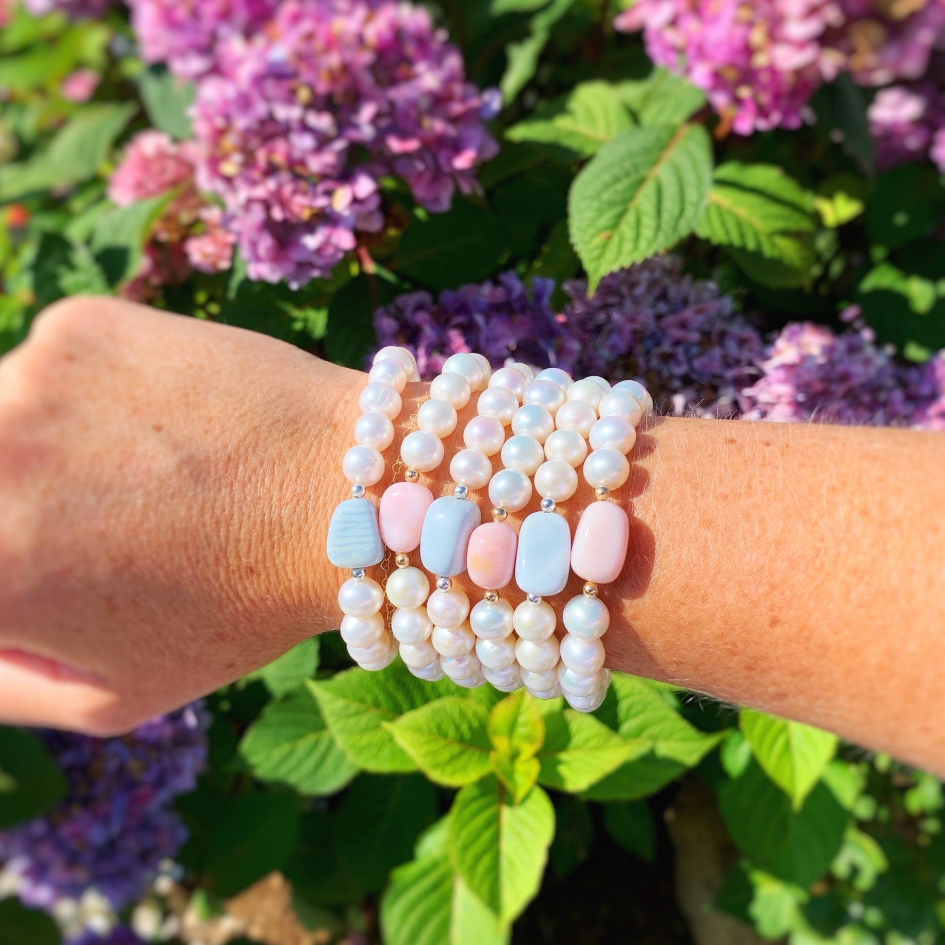 pink peony and blue hydrangea bracelets are pictured here worn on a wrist in an every other pattern of blue/pink with 6 bracelets. the bracelets feature either a pink or blue opal at the center with freshwater pearls on the rest. this picture has all of the bracelets on a wrist with pink hydrangeas in the background