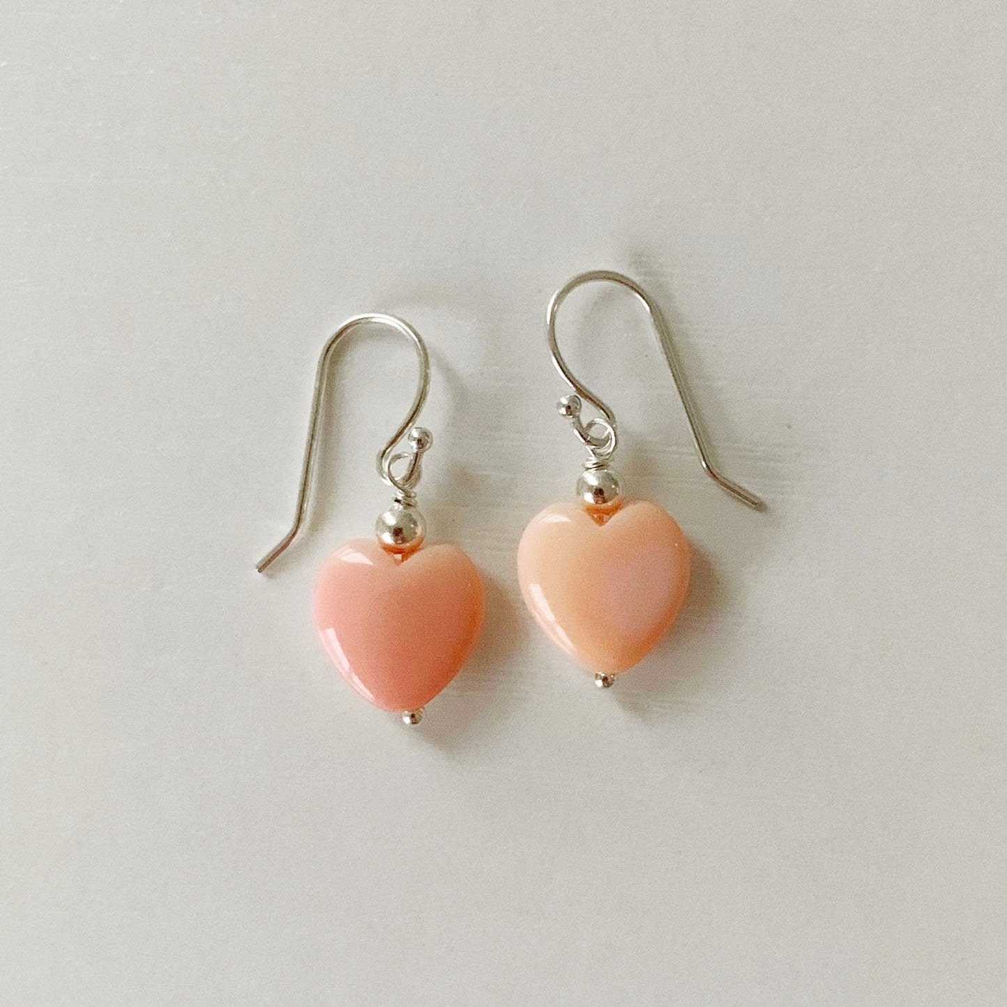 the pink posy earrings by mermaids and madeleines are simple drop earrings created with sterling beads and findings and feature a conch shells carved into hearts. this pair is photographedon a white surface