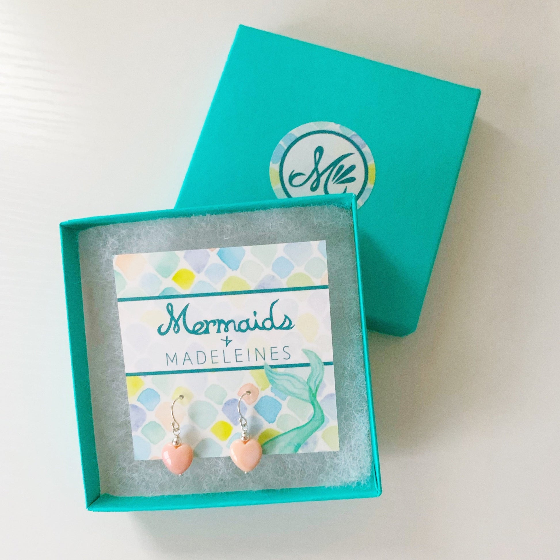 the pink posy earrings by mermaids and madeleines are simple drop earrings created with sterling silver beads and findings and feature a conch shells carved into hearts. this pair is carded and photographed in a teal gift box on a white surface