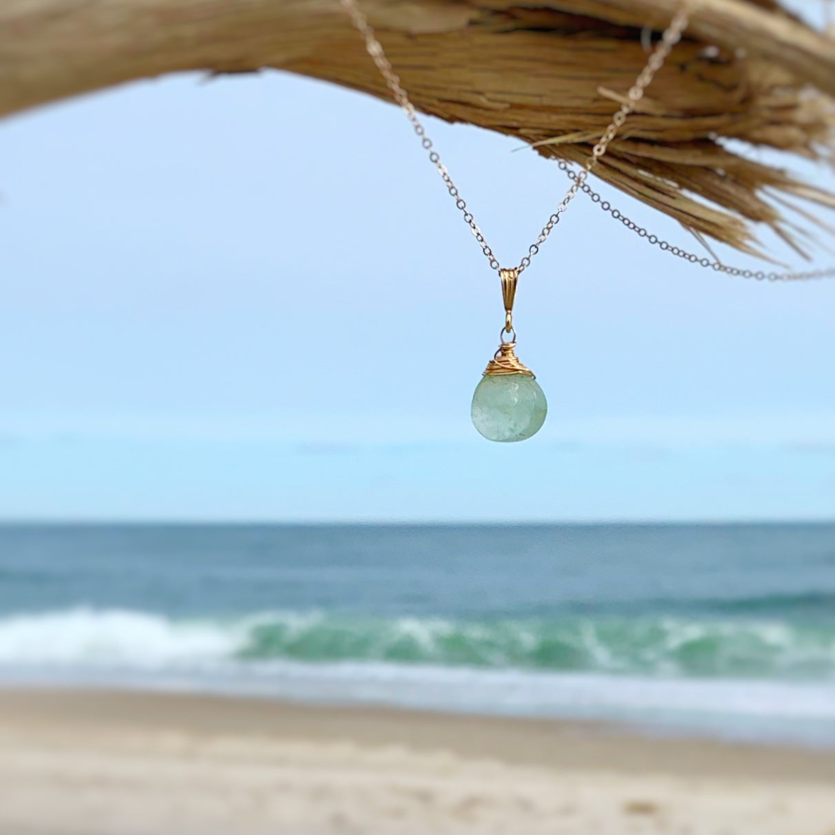 the raindrop necklace by mermaids and madeleines is a pendant style necklace featuring a faceted briolette of natural color aquamarine, wire wrapped in 14k gold filled wire and hanging from a dainty 14k gold filled chain. this necklace is pictured on a piece of drift wood on the beach