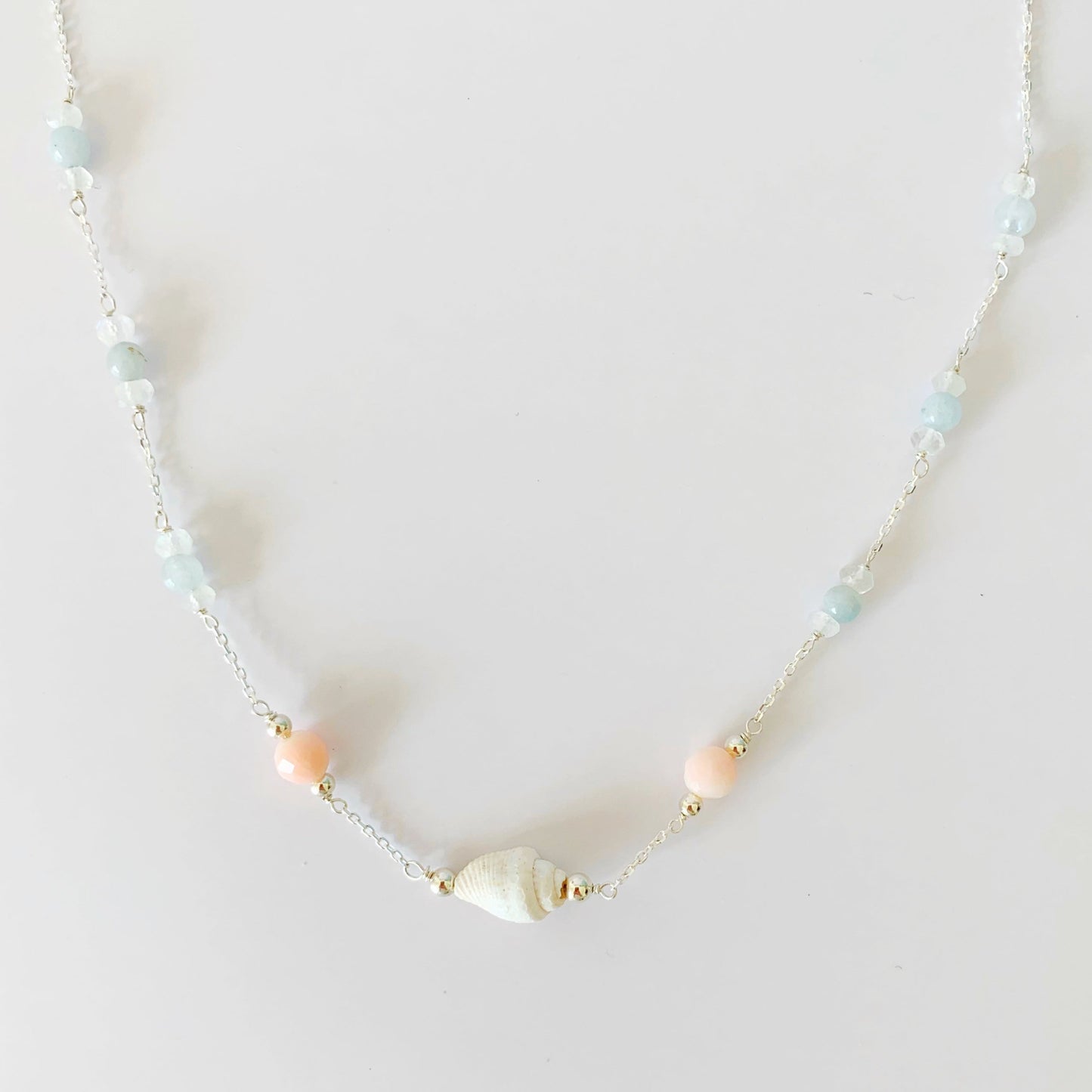 the seersucker necklace by mermaids and madeleines is a station style necklace with a shell and coral beads at the center with segments of moonstone and aquamarine along the sterling chain to the clasp. this necklace is photographed on a white surface