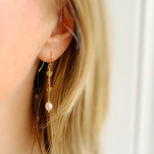 the sun and snow earrings by mermaid and madeleines are a simple drop earrings on 14k gold filled chain that feature 2 faceted citrine beads with a freshwater drop pearl at the bottom of the earring. this is an image of the earrings being worn on an ear and viewed close up