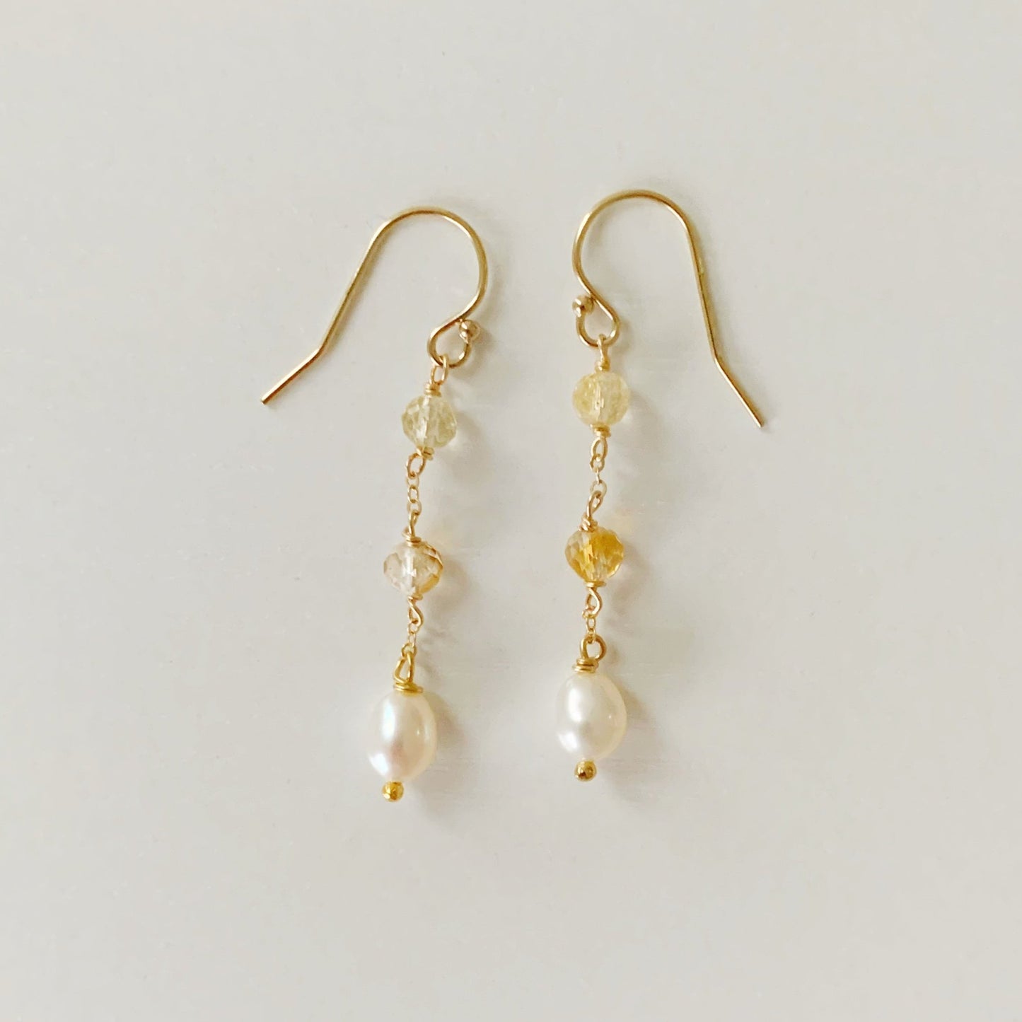 the sun and snow earrings by mermaid and madeleines are a simple drop earrings on 14k gold filled chain that feature 2 faceted citrine beads with a freshwater drop pearl at the bottom of the earring. this is an image of the earrings photographed flat on a white surface