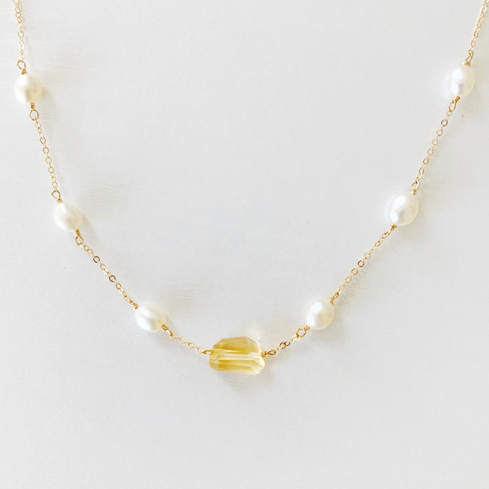 the sun and snow neckklace by mermaids and madeleines is a delicate station necklace with a citrine gem at the center and freshwater rice pearls going up the chain the 14k gold filled chain. this necklace  is viewed closer up on a flat white surface