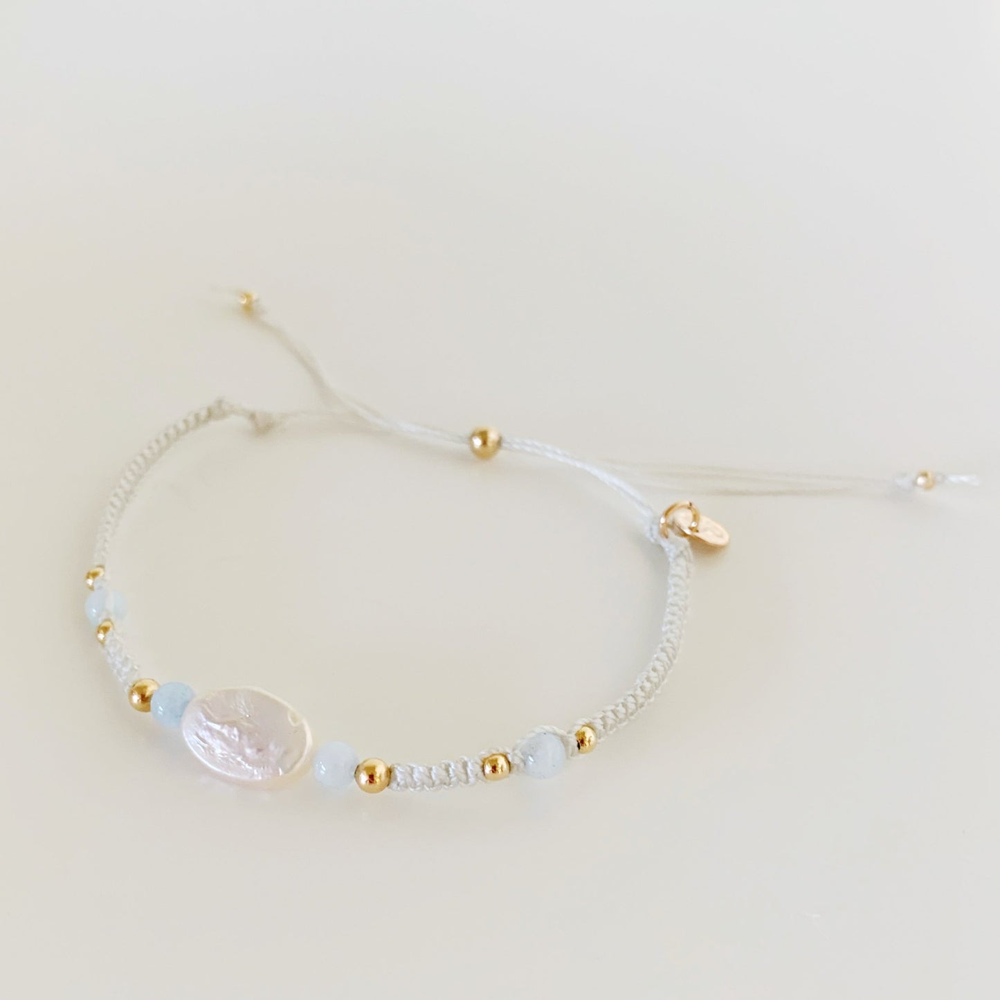 the world is your oyster bracelet by mermaids and madeleines is an adjustable macrame style bracelet created with white shell cord with a freshwater oval pearl at the center with aquamarine beads on either side. this braclet is photographed from the top down on a white surface