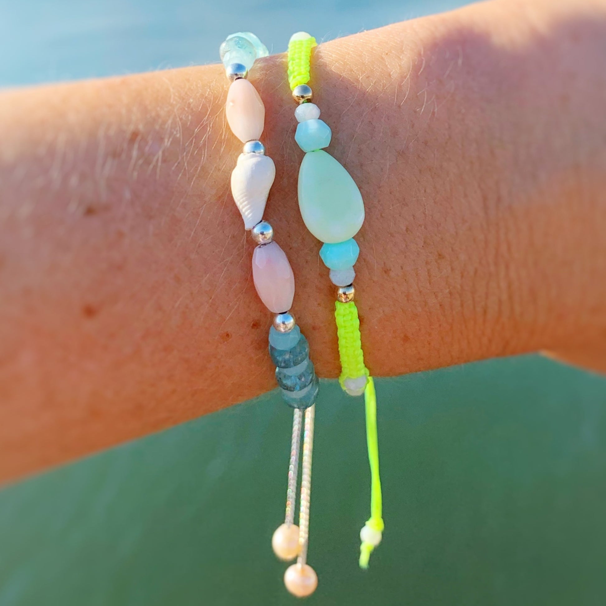 Two bracelets pictured worn on a wrist over an ocean background. The bracelets are a shellebration bracelet that features alternating peruvian opal and apatite beads with a shell at center and a Captiva macrame bracelet in neon yellow