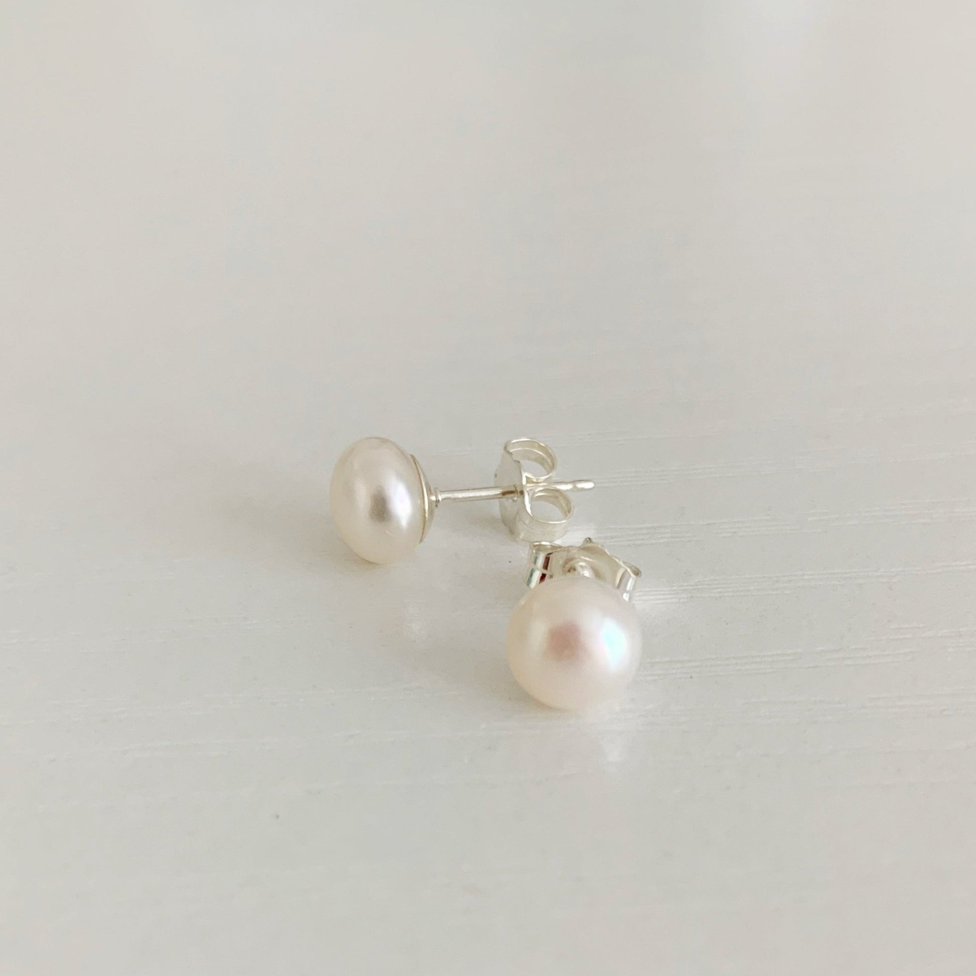 Pearlfect Day pearl earrings by mermaids and madeleines feature freshwater button pearls with sterling silver post earrings. this pair is photographed with one facing forward to the camera and the other to the side to show multiple angles of the post earrings. this picture is taken on a white surface